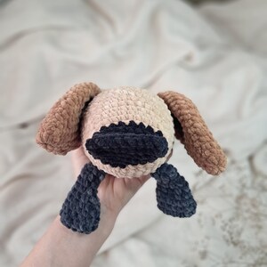 May include: A handmade crochet dog toy in beige, brown, and navy blue. The dog has long floppy ears, a dark blue snout, and dark blue feet. The toy is soft and plush, perfect for a child.