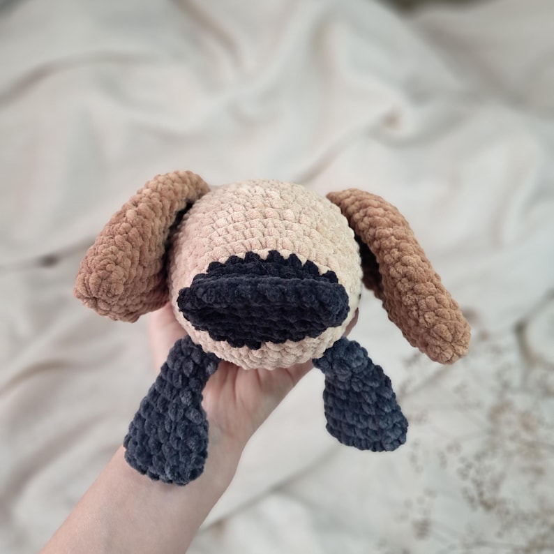 May include: A handmade crochet dog toy in beige, brown, and navy blue. The dog has long floppy ears, a dark blue snout, and dark blue feet. The toy is soft and plush, perfect for a child.