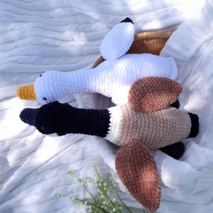 May include: Two plush goose toys. One is white with a yellow beak and black accents, the other is brown and black with tan accents. The toys are made of a soft, textured material and are lying on a white blanket.