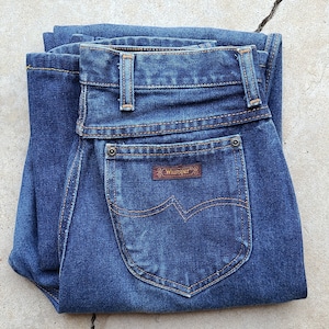 Puede incluir: Un par de pantalones vaqueros azules de mezclilla doblados por la mitad, con un parche de cuero marr&oacute;n que dice "Wrangler" en el bolsillo trasero.