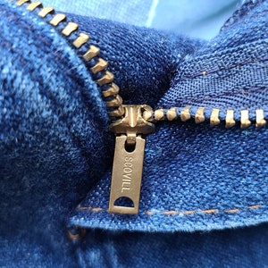 Puede incluir: Primer plano de una cremallera de mezclilla azul con un tirador de metal dorado. El tirador de la cremallera tiene la palabra "SCOVILL" grabada.