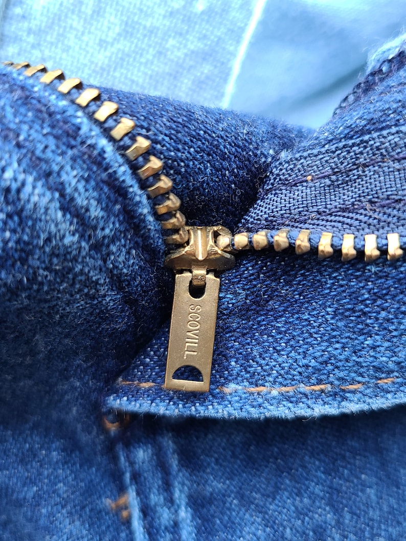 Puede incluir: Primer plano de una cremallera de mezclilla azul con un tirador de metal dorado. El tirador de la cremallera tiene la palabra "SCOVILL" grabada.