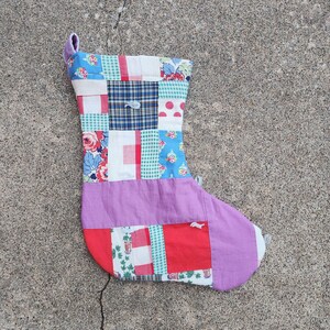 May include: A patchwork Christmas stocking with a purple, red, blue, green, and white color scheme. The stocking is made of fabric scraps sewn together in a random pattern.