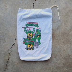 May include: White canvas drawstring bag with a Teenage Mutant Ninja Turtles graphic. The graphic features three turtles in action poses with the words "Teenage Mutant Ninja Turtles" and "Turtles" printed above them.