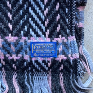 Może przedstawiać: Niebieski i biały koc Pendleton z wełny z r&oacute;żowo-czarnym wzorem w kratę. Koc ma frędzle na krawędziach i niebieską metkę z napisem "WARRANTED TO BE A PENDLETON" i "PURE VIRGIN WOOL MADE IN USA".