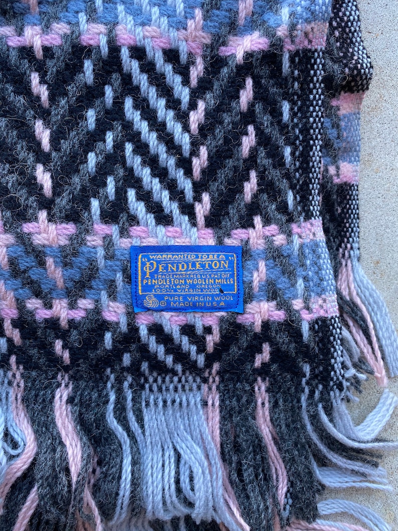 Może przedstawiać: Niebieski i biały koc Pendleton z wełny z r&oacute;żowo-czarnym wzorem w kratę. Koc ma frędzle na krawędziach i niebieską metkę z napisem "WARRANTED TO BE A PENDLETON" i "PURE VIRGIN WOOL MADE IN USA".