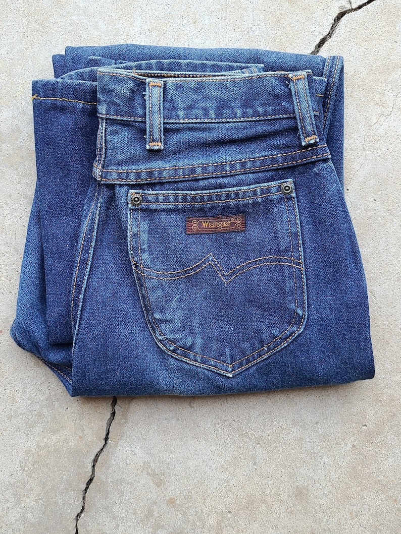 Puede incluir: Un par de pantalones vaqueros azules de mezclilla doblados por la mitad, con un parche de cuero marr&oacute;n que dice "Wrangler" en el bolsillo trasero.