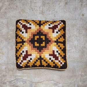 May include: A square-shaped cushion with a geometric pattern in shades of brown, yellow, and white. The cushion is made of a soft, textured fabric.