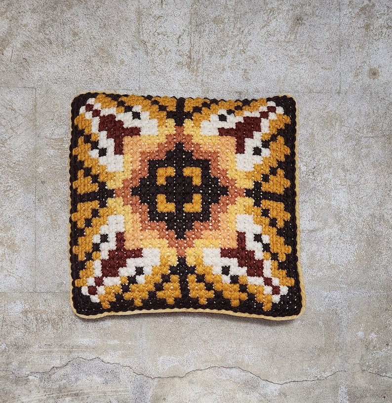May include: A square-shaped cushion with a geometric pattern in shades of brown, yellow, and white. The cushion is made of a soft, textured fabric.