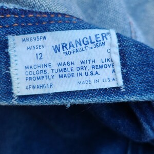 Puede incluir: Una etiqueta blanca Wrangler 'No-Fault' Jean con costuras azules. La etiqueta tiene el texto 'MR695PW Misses 12' y 'Machine Wash With Like Colors, Tumble Dry, Remove Promptly Made in U.S.A. 4FWAH61R Made in U.S.A.'