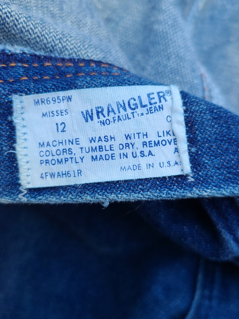 Puede incluir: Una etiqueta blanca Wrangler 'No-Fault' Jean con costuras azules. La etiqueta tiene el texto 'MR695PW Misses 12' y 'Machine Wash With Like Colors, Tumble Dry, Remove Promptly Made in U.S.A. 4FWAH61R Made in U.S.A.'