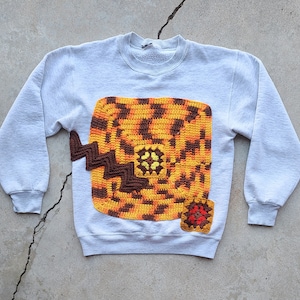 May include: A gray sweatshirt with a brown, orange, and yellow crocheted design on the front. The design is a square with a smaller square in the center.