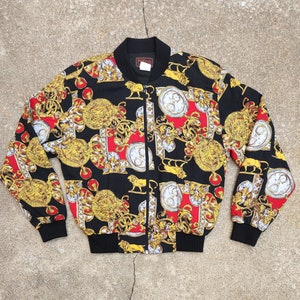 May include: A black bomber jacket with a gold and red patterned design featuring a repeating motif of ornate clocks and lions. The jacket has a zipper closure and a stand-up collar. The label on the jacket reads "Polo".