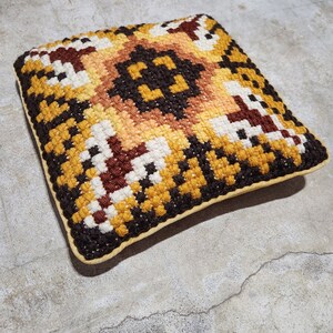 May include: A square-shaped cushion with a geometric pattern in shades of brown, yellow, and white. The cushion has a brown border and a cream-coloured backing.