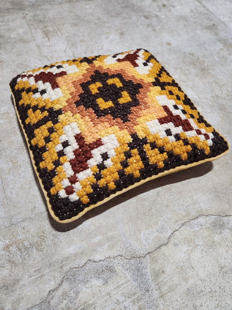 May include: A square-shaped cushion with a geometric pattern in shades of brown, yellow, and white. The cushion has a brown border and a cream-coloured backing.