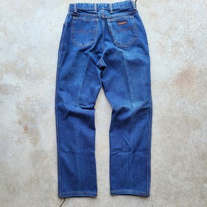 Puede incluir: Un par de pantalones vaqueros de mezclilla azul oscuro con un parche de cuero marr&oacute;n en el bolsillo trasero que dice "Wrangler".