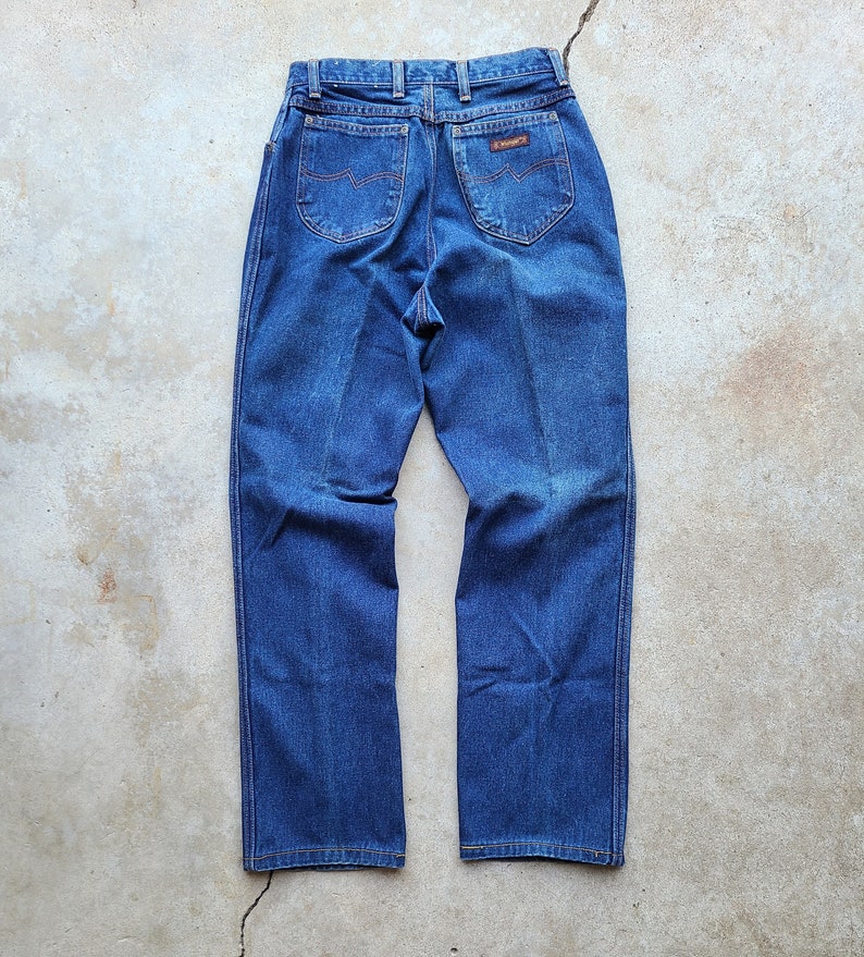 Puede incluir: Un par de pantalones vaqueros de mezclilla azul oscuro con un parche de cuero marr&oacute;n en el bolsillo trasero que dice "Wrangler".