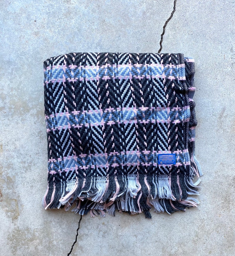 Może przedstawiać: Czarny, r&oacute;żowy i niebieski pled z frędzlami. Pled ma tkane logo z napisem "Pendleton".