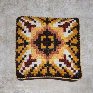 May include: A square cushion with a geometric pattern in shades of brown, yellow, and white. The cushion is made of a woven fabric and has a tufted texture.