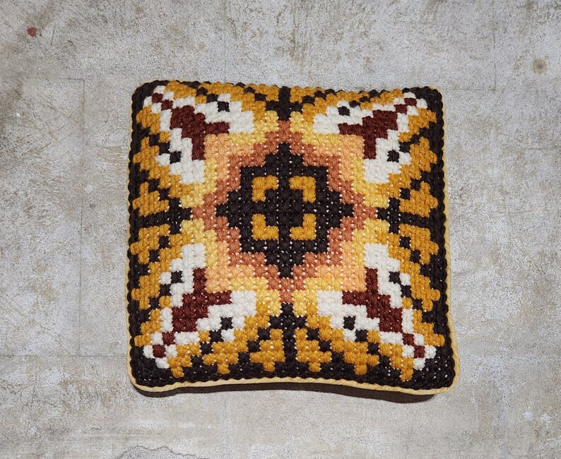 May include: A square cushion with a geometric pattern in shades of brown, yellow, and white. The cushion is made of a woven fabric and has a tufted texture.