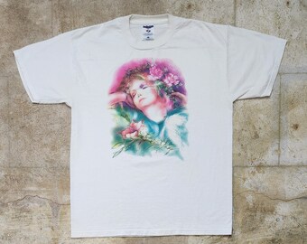May include: White t-shirt featuring a colourful image of a woman with flowers in her hair. The image is in a watercolour style.