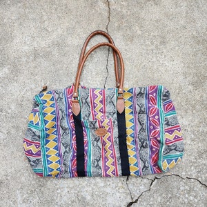 May include: A colorful travel duffel bag with a geometric pattern in shades of purple, yellow, blue, and green. The bag has brown leather straps and a zipper closure.