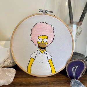 May include: Embroidered hoop art featuring a yellow cartoon character with a pink afro, wearing a white shirt. The character is smiling with a brown mouth and yellow eyes.