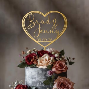 Op de afbeelding: Een goudkleurige taarttopper in de vorm van een hart met een gelaagde omtrek. In het midden staan de namen Brad & Jenny in een cursief lettertype, met daaronder de datum 09.09.26. De topper is in een taart met rozen gestoken.