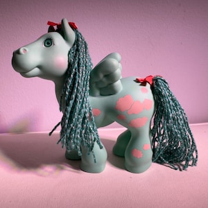 May include: A vintage teal-colored toy pony with pink cloud-shaped markings on its flank. The pony has blue braided hair, wings, and a red bow on its head and tail. The toy is a collectible item.