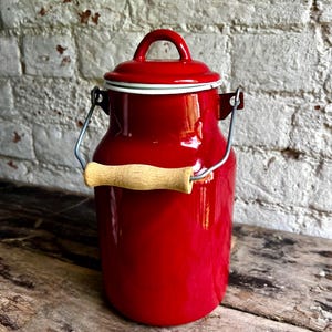 May include: A vibrant red enamel milk churn with a matching lid and a wooden handle. It features a silver wire handle and a white trim around the lid. The churn is on a wooden surface.