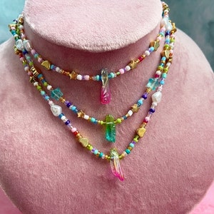 Op de afbeelding: Een gelaagde ketting met kleurrijke kralen, gouden sterren en hartcharms. De ketting heeft drie hangende kristal-achtige bedels in de kleuren roze, groen en blauw.