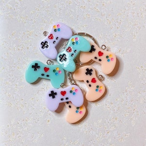 May include: A set of six colorful plastic keychains shaped like video game controllers. The keychains are in shades of light blue, lavender, and peach. Each keychain has a small heart and colorful buttons.