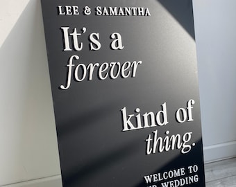 May include: A black wedding sign with white raised lettering. The sign reads "LEE & SAMANTHA" at the top, followed by "It's a forever kind of thing." and "WELCOME TO OUR WEDDING" at the bottom.