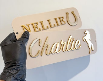 Può includere: Due cartelli in legno beige con lettere dorate. Il cartello superiore dice "Nellie" con un ferro di cavallo dorato. Il cartello inferiore dice "Charlie" con una silhouette bianca di un cavallo.