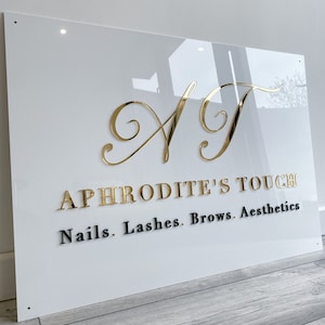 May include: White acrylic sign with gold lettering that reads "A.T. Aphrodite's Touch" and black lettering that reads "Nails. Lashes. Brows. Aesthetics."