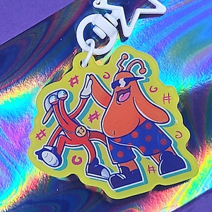 May include: A yellow acrylic keychain featuring two cartoon characters giving a high-five. One character is orange with sunglasses and blue polka-dot shorts. The other is red. The keychain has a white star-shaped clasp.
