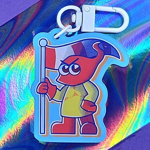 Puede incluir: Un llavero de dibujos animados con un personaje rojo sosteniendo una bandera con rayas rojas, blancas y azules. El personaje tiene una expresión enfadada y lleva una camisa amarilla y zapatos rojos. El llavero tiene un cierre blanco.