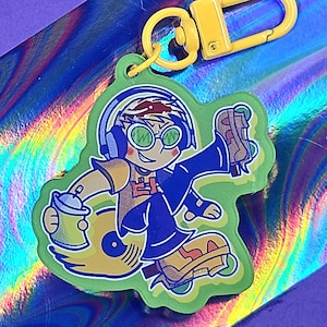 May include: A green acrylic keychain featuring a cartoon character with headphones, green glasses, and a spray paint can. The character is on roller skates and wearing a blue jacket. The keychain has a yellow clasp.