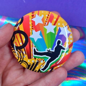 May include: A circular pin-back button featuring a vibrant yellow background with red and orange vertical stripes, black circular outlines, and white polka dots. A black silhouette of a person jumping is centered over colorful star bursts.