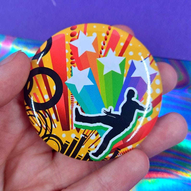 May include: A circular pin-back button featuring a vibrant yellow background with red and orange vertical stripes, black circular outlines, and white polka dots. A black silhouette of a person jumping is centered over colorful star bursts.