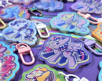 Pode incluir: Uma coleção de chaveiros de pôneis de desenho animado coloridos com vários designs e clipes de mosquetão anexados. Os chaveiros apresentam diferentes pôneis em tons de roxo, rosa, amarelo e azul, com um fundo holográfico.