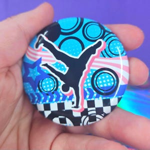 May include: A circular pin-back button featuring a black silhouette of a person performing a handstand. The background displays a vibrant pattern of blue circles, pink wavy stripes, a blue star, and a black and white checkered border.