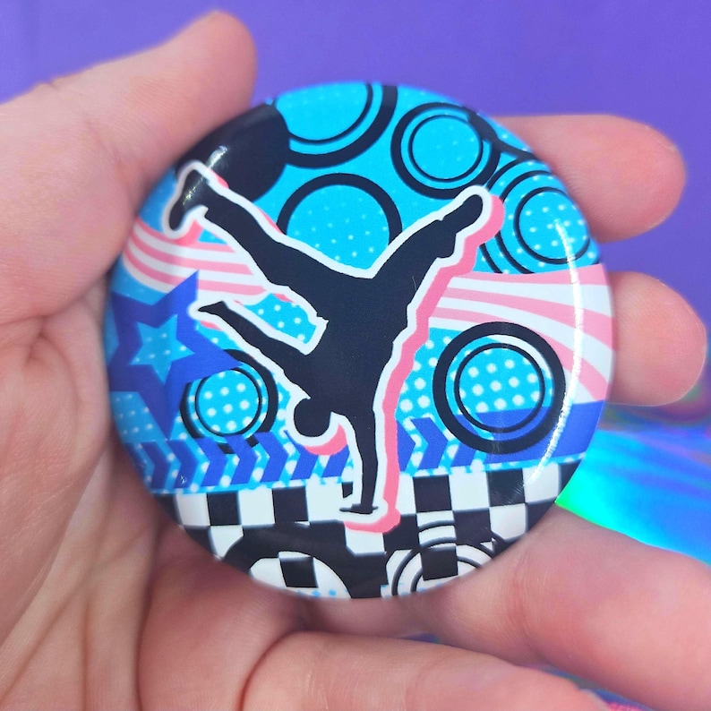 May include: A circular pin-back button featuring a black silhouette of a person performing a handstand. The background displays a vibrant pattern of blue circles, pink wavy stripes, a blue star, and a black and white checkered border.