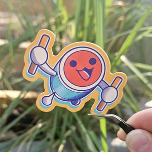 May include: A colorful sticker featuring a cartoon character with a red face, blue body, and orange outline. The character has a happy expression and is holding two drumsticks. The sticker is being held by tweezers.