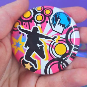 May include: A circular pin-back button featuring a black silhouette of a person jumping against a vibrant background of pink and white sunburst rays, yellow stars, black concentric circles, and a blue globe icon with a white hand cursor.