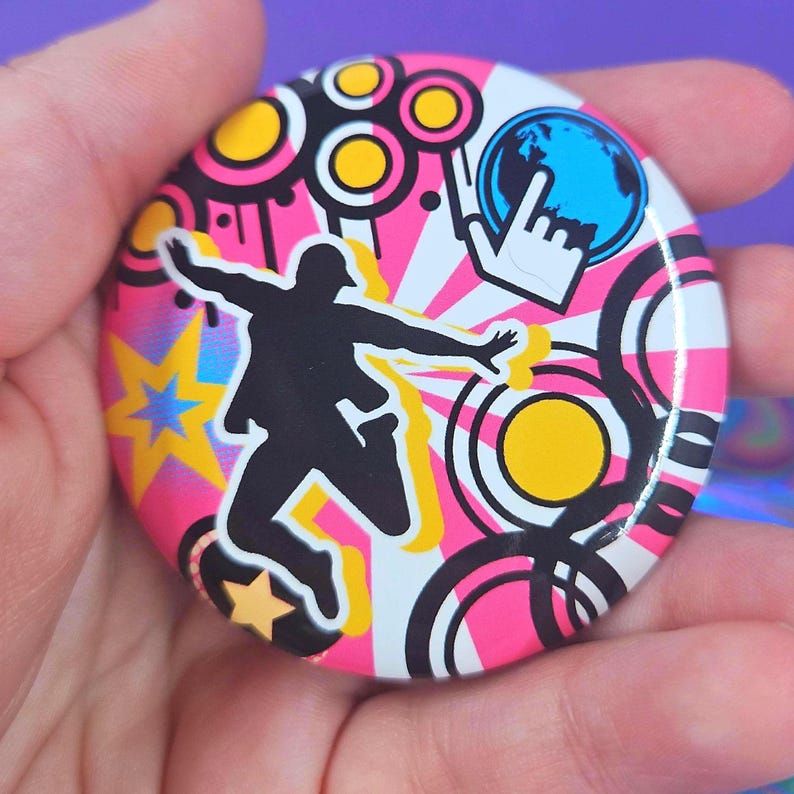 May include: A circular pin-back button featuring a black silhouette of a person jumping against a vibrant background of pink and white sunburst rays, yellow stars, black concentric circles, and a blue globe icon with a white hand cursor.