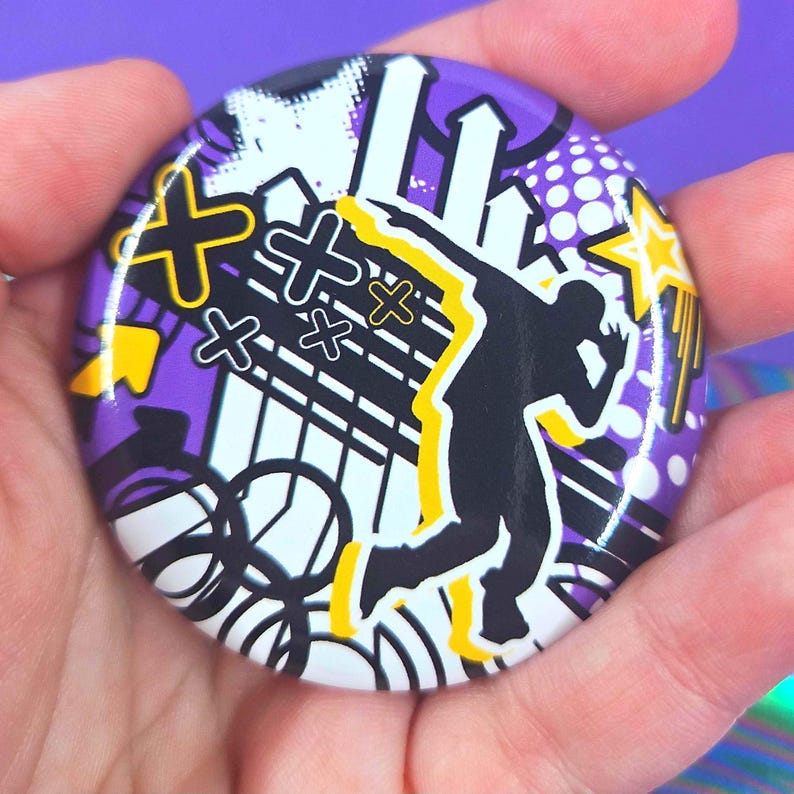May include: A circular pin-back button featuring a vibrant graphic design with a purple background. The artwork displays a black silhouette of a person dancing, accented by yellow outlines, white arrows, black circular patterns, and yellow star shapes.