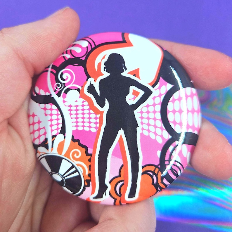 May include: A circular pin-back button featuring a black silhouette of a person standing with one hand on their hip. The background is a vibrant graphic design with pink, orange, white, and black patterns including swirls, dots, and bold shapes.