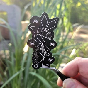 May include: Black sticker with a white geometric design, held by tweezers. The design includes diamond shapes, starbursts, and Japanese text. The sticker is held in front of a blurred green background.