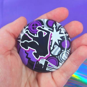May include: A circular pinback button featuring a black silhouette of a person jumping against a background of purple, white, and black geometric shapes, including a starburst and circular target patterns with a glossy finish.